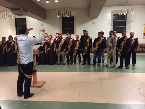 Music instructor conducts a choir wearing black robes with gold trim performing in an indoor venue